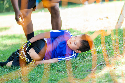 Young Hispanic boy holds his knee while laying on the soccer field. He has injured his knee. He is wearing protective gloves and a blue jersey. An African American coach or referee is talking with him. His face grimmaces in pain.