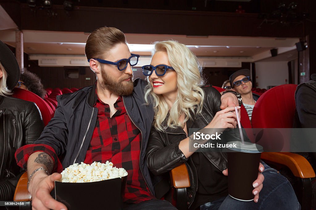 Young couple in 3D movie theater Happy young couple wearing 3D glasses, sitting in the cinema and watching 3D movie. Bearded young man holding popcorn, blonde woman holding drink. 3-D Glasses Stock Photo