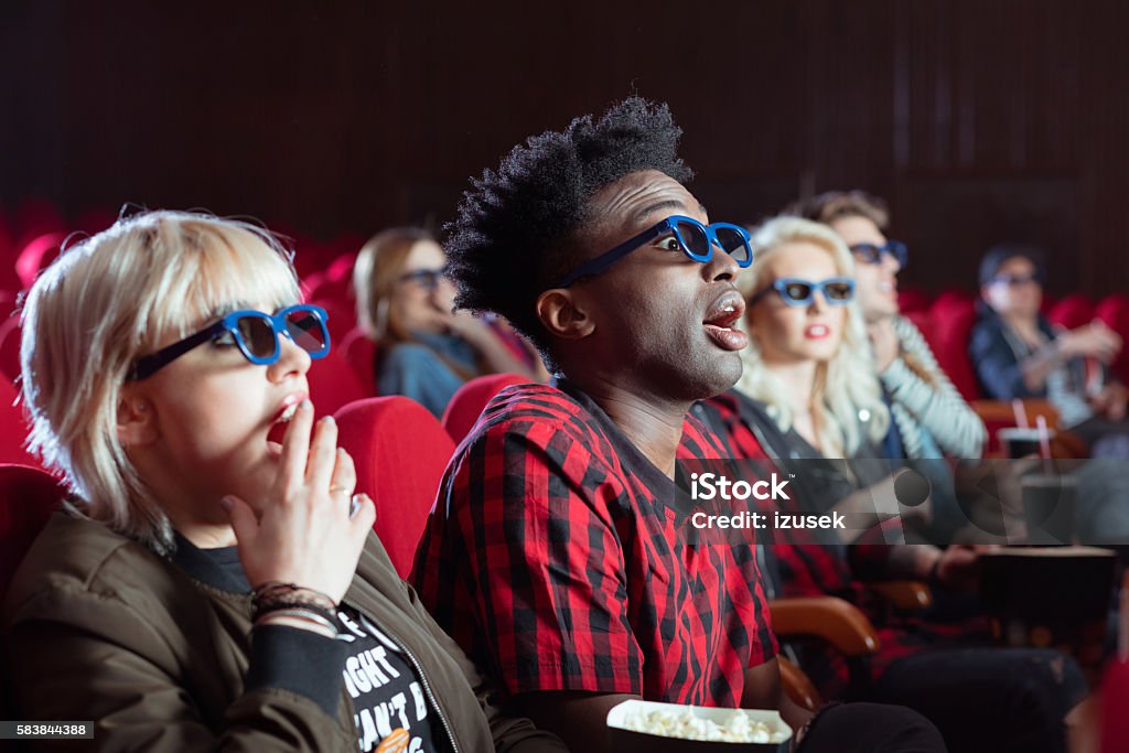 Young people in 3D movie theater Multi ethnic group of young adult people wearing 3D glasses, sitting in the cinema and watching scary 3D movie. Focus on afro american young man. Human Face Stock Photo