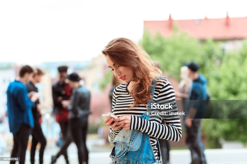 Young woman using smart phone outdoor Young woman texting on smart phone outdoor. Group of young people in the background. Mobile Game Stock Photo