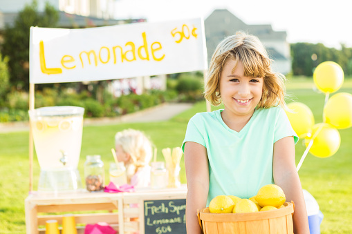 Pretty little girl with blonde hair wearing a mint colored shirt is holding up a basket full of bright yellow lemons. She is smiling directly at the camera. There is a lemonade stand behind her with a sign that says \