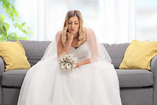 Depressed blond bride sitting on a sofa and looking down at home
