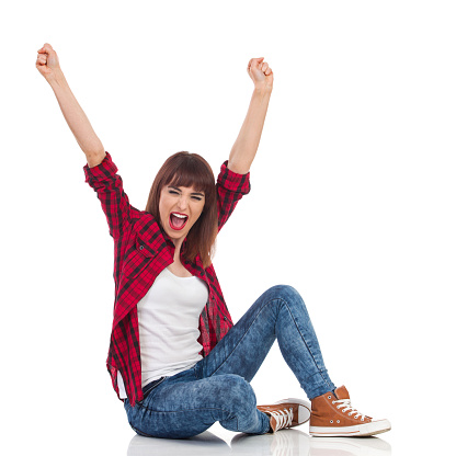 Excited young woman in red lumberjack shirt, jeans and brown sneakers sitting on a floor with arms raised and shouting. Full length studio shot isolated on white.
