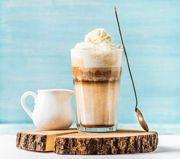 Latte macchiato with whipped cream, serving silver spoon and pitcher Latte macchiato with whipped cream, serving silver spoon and pitcher on wooden round board over blue painted wall background, selective focus, horizontal composition mocha stock pictures, royalty-free photos & images