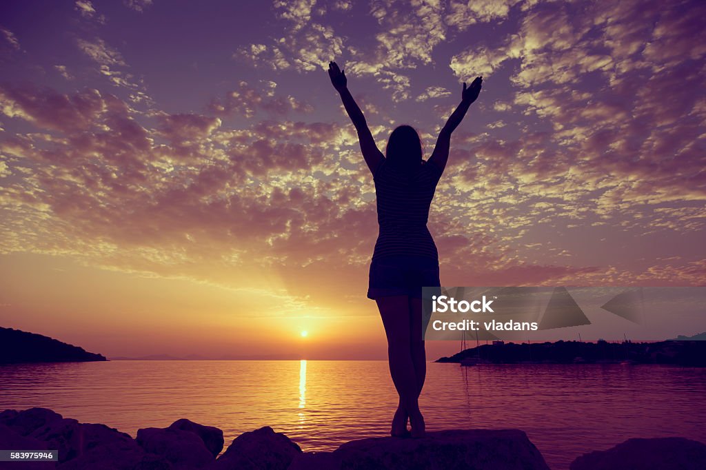 Freedom Young woman standing on a rocky shore of Syvota bay, Greece, looking at sunset and enjoying beauty and freedom Adult Stock Photo