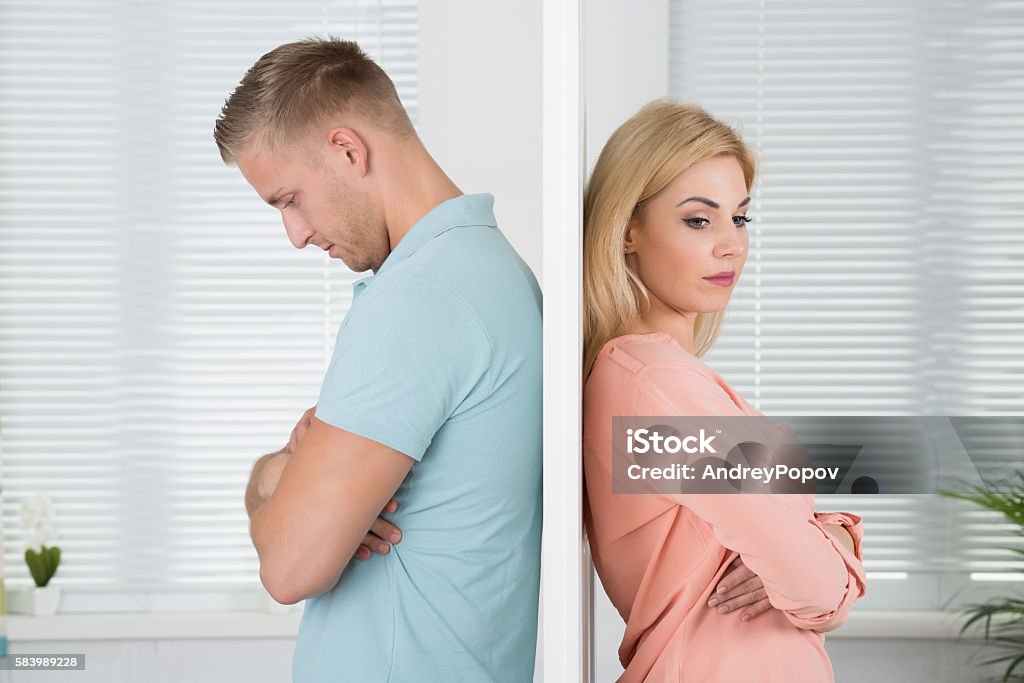 Unhappy Couple Standing Back To Back At Home Side view of unhappy young couple standing back to back at home Divorce Stock Photo