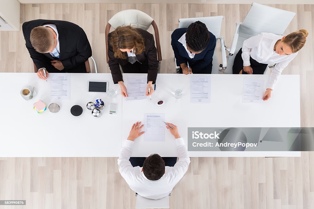 Businesspeople Interviewing Young Male Applicant High Angle View Of Businesspeople Interviewing Young Male Applicant In Office Interview - Event Stock Photo