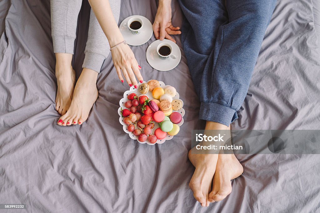 Young caucasian couple having romantic breakfast in bed Young caucasian couple having romantic breakfast in bed. Closeup of female and male feet, two cups of coffee, fruits and colorful biscuits. Eating Stock Photo