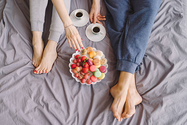 Young caucasian couple having romantic breakfast in bed Young caucasian couple having romantic breakfast in bed. Closeup of female and male feet, two cups of coffee, fruits and colorful biscuits. bed human foot couple two parent family stock pictures, royalty-free photos & images