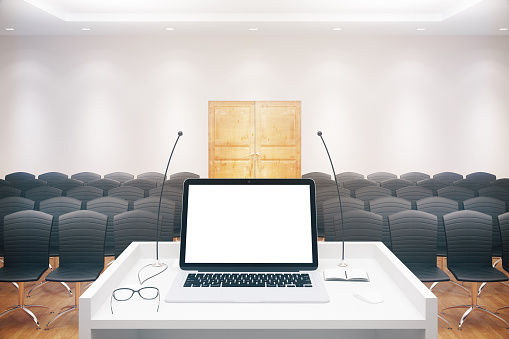 Blank white laptop placed on speaker's stand in conference hall interior with rows of seats and wooden door. Mock up, 3D Rendering