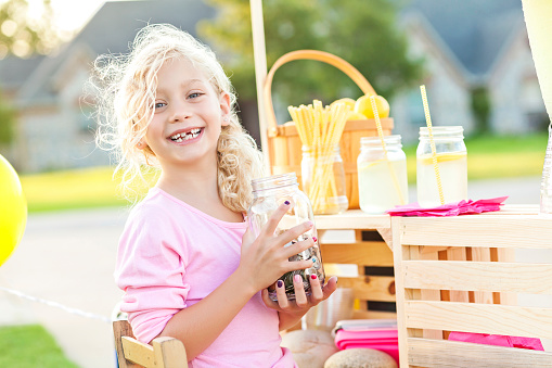 Adorable little girl with curly blond hair. She is smiling a big toothy smile at the camera. She has a jar of coins and cash that she has earned while selling lemonade. There are cups of lemonade and straws behind her. There is even a basket full to the top of lemons. The little girl is alone, she is wearing a light pink shirt.