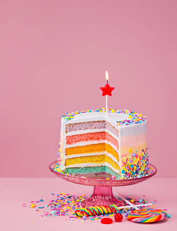 Colorful layered Birthday cake with sprinkles and candy over a pink background.