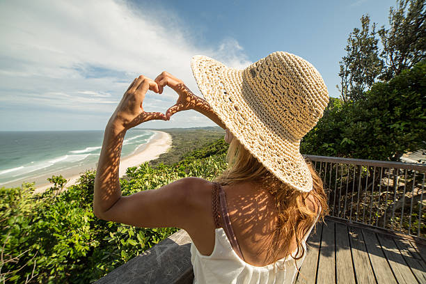 Caucasian woman makes a heart shape finger frame above Tallow beach in Byron Bay, Australia.