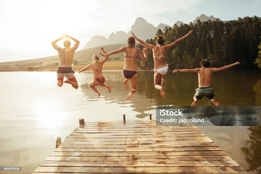 Young friends jumping into lake from a jetty Portrait of young friends jumping from jetty into lake. Friends in mid air on a sunny day at the lake. Lake Stock Photo