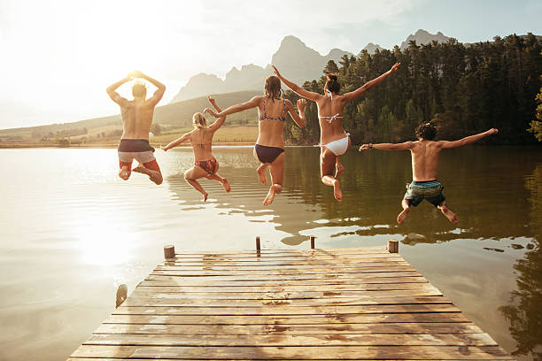 Young friends jumping into lake from a jetty Portrait of young friends jumping from jetty into lake. Friends in mid air on a sunny day at the lake. jetty stock pictures, royalty-free photos & images