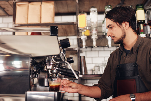Side view of male barista preparing espresso at coffee shop. Young man in apron making coffee using coffee maker at the cafe.