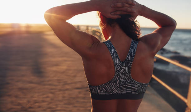 Muscular woman in sports bra at sunset Rear view shot of muscular woman in sports bra standing outdoors with her hands on her head at sunset. sports bra stock pictures, royalty-free photos & images