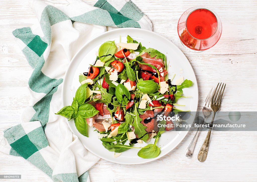 Summer arugula, prosciutto, strawberry salad with glass of rose wine Summer arugula, prosciutto and strawberry salad with glass of rose wine over white painted wooden background, top view, horizontal composition Salad Stock Photo
