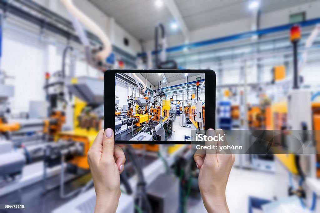 Ordering on-line from plastic factory Horizontal color image of female hands holding a digital tablet in a modern plastic production line. Ordering on-line from injection moulding factory on a touchscreen tablet computer. Large factory, industrial machines, robots and manufacturing equipment arranged in background. Manufacturing Stock Photo