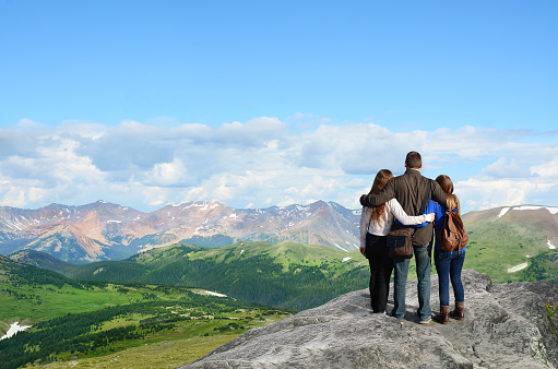 Family on hiking trip in  Rocky Mountains National Park, Colorado, USA. Father with arms around his family looking at beautiful summer mountains landscape. People standing on  top of  mountain rock. 
