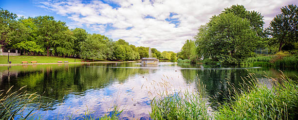 Panoramic view of Montreal's Parc Lafontaine, featuring the large pond, active fountain, reeds and trees. The partloud cloudy sky is nicely reflected on the water.