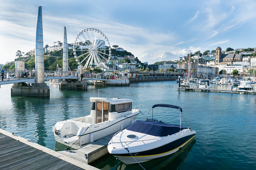 Torquay,England - July 22, 2016: Torquay is a major town and tourist destination in south Devon and is withinTorbay. People can be seen on the harbour walkway, two pleasure boats are moored in the foreground and the big wheel is in action. It is a sunny day in summer.