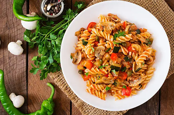 Vegetarian Vegetable pasta Fusilli with zucchini, mushrooms and capers in white bowl on wooden table. Top view