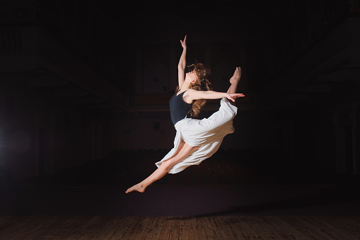 Photo of young brunette dancer girl, ballerina in white skirt in split jump on stage in theater with spotlight. barefoot dancer.