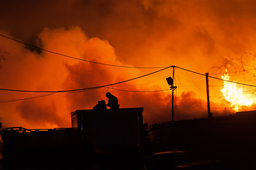 Firefighters battles storage fire. Silhouette of firefighters on the roof. Night Scene with a fiery glow in the background.