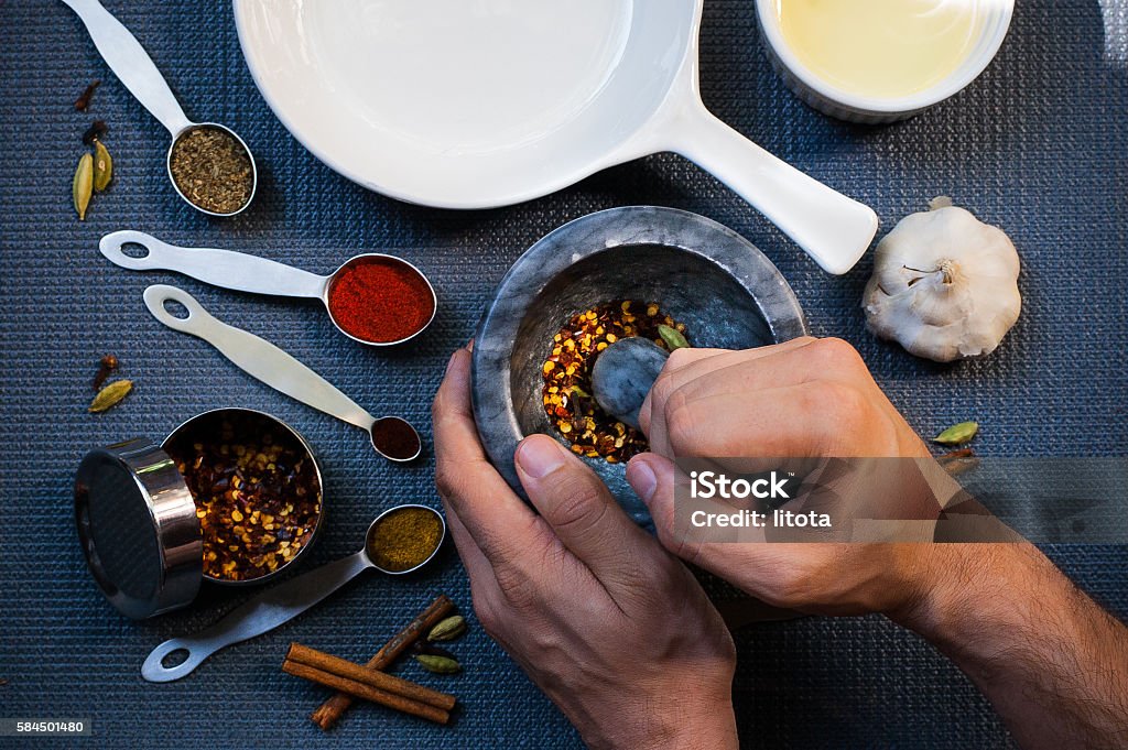 One man grinding Spices A man is grinding spices with a mortar and pestle. Kitchenware and other spices (oregano, paprika, red pepper, black pepper, cinnamon sticks, cardamon, cloves) are placed at the table. Taken on 7.29.2016 in the US, Portland, OR. Mortar and Pestle Stock Photo