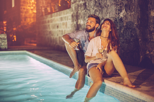 Romantic couple having aperitif near the swimming pool. Sitting at poolside by night, having fun.