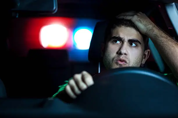 Police in pursuit of a reckless driver at night. About 30 years old Caucasian man, worried and holding his head, looking back at a police car.
