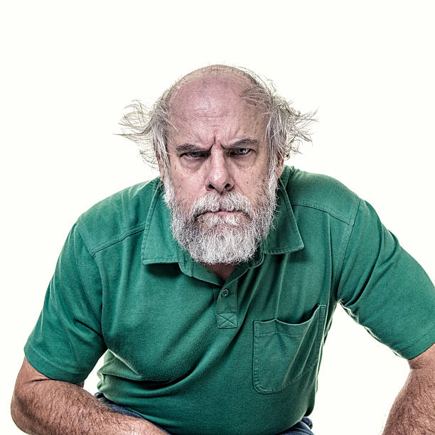 An angry, stressed, menacing, overworked, balding senior adult businessman with tangled grey hair is staring at the camera with a humorous, exaggerated green eyed facial expression. He has a scruffy, full beard and mustache. He is casually dressed wearing a green pullover polo shirt with a collar and a front chest pocket.