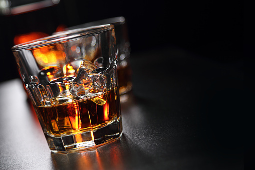 Two glasses of whiskey with ice cubes, and bottle in background, on the bar. Glasses reflected on the surface of the bar. Focus on front glass and blurred dark background. Close-up photography. Horizontal orientation.