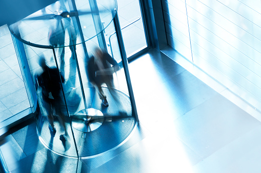 A high angle view of a people entering and exiting an office building through a revolving door. A slow shutter speed allows for the anonymity of the people as they quickly move through the rotating door.  A cool blue cast dominates the scene.