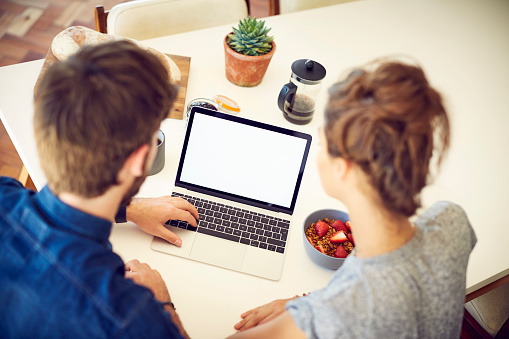 High angle over the shoulder view of couple using laptop with blank screen at home. Young male and female partners sitting at dining table. Rear view of bloggers are at home.