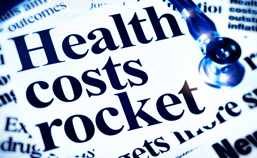 A stethoscope sits on newspaper headlines dealing with rising healthcare costs. Blue toned.