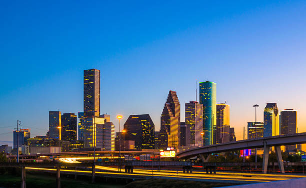 Downtown Houston Skyline at Sunset with Orange Reflection Downtown Houston skyline at sunset / dusk with a blue and orange sky, orange reflection on buildings from the sunset, and a freeway / highway with light trails. houston skyline stock pictures, royalty-free photos & images
