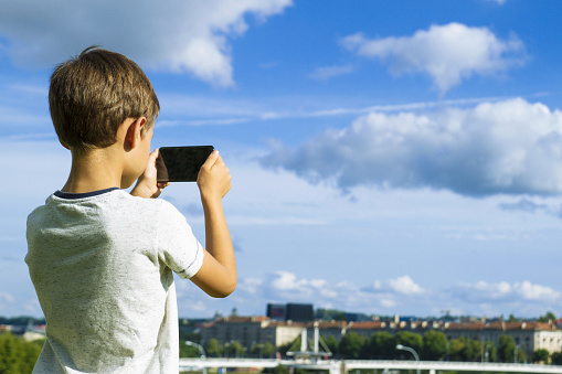 Boy with mobile phone. Child taking photo with his smartphone. Beautiful sky and city background. Back view. Technology, leisure, gaming concept