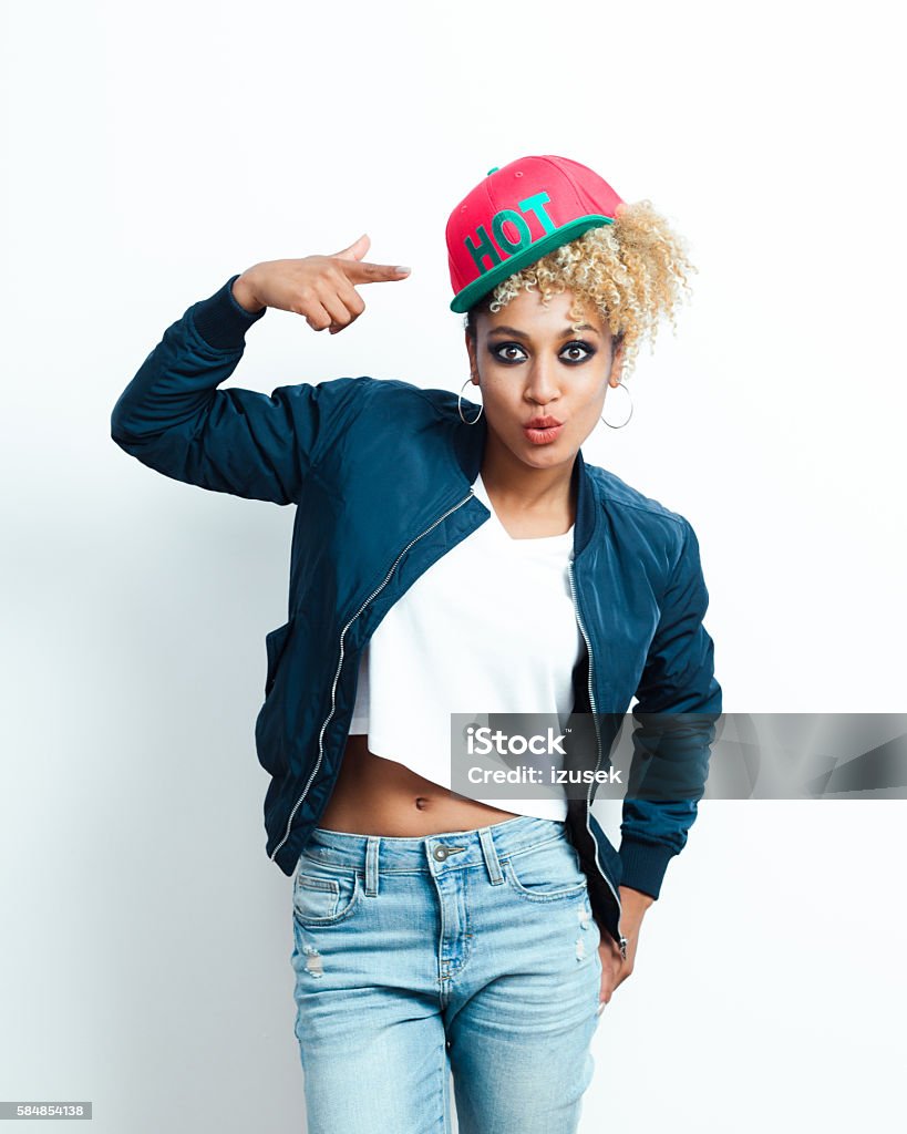 Portrait of afro american young woman with pink cap Portrait of afro american young woman wearing bomber jacket, denim trausers and pink baseball cap, pointing at cap with index finger. Jacket Stock Photo