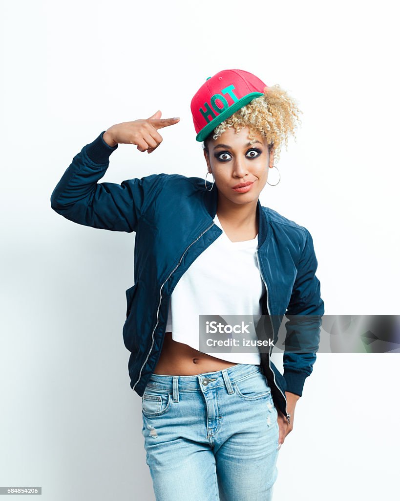 Portrait of afro american young woman with pink cap Portrait of surprised afro american young woman wearing bomber jacket, denim trausers and pink baseball cap, pointing at cap with index finger. Adult Stock Photo