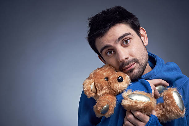 Nostalgia Young at Heart. Cute young man embracing stuffed toy and looking at camera. Black-haired man remembering his childhood. He has emotion like child. Its photo illustrating infant behavior. It is perfect for using it in commercial and advertising photography, reports, books, presentation man with teddy bear stock pictures, royalty-free photos & images