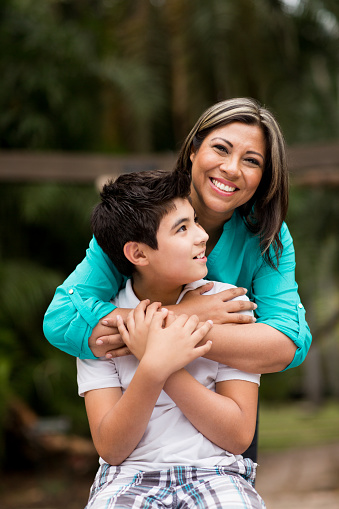 A cheerful latin mother and her teen son embracing and smiling in a vertical medium shot outdoors.