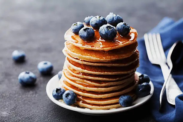 Stack of baked american pancakes or fritters with blueberries and honey syrup on rustic black table. Delicious dessert for breakfast.