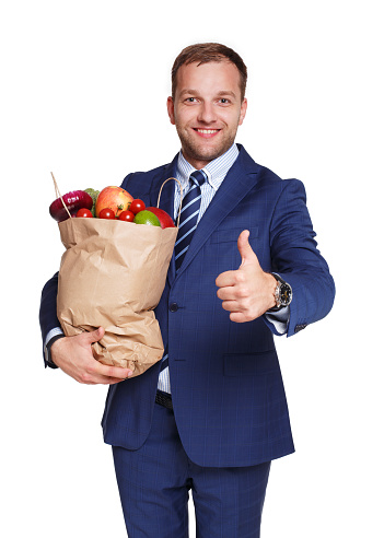 Smiling young businessman hold shopping bag full of groceries isolated at white background. Healthy food shopping. Paper package with vegetables and fruits, happy man buyer show thumb up