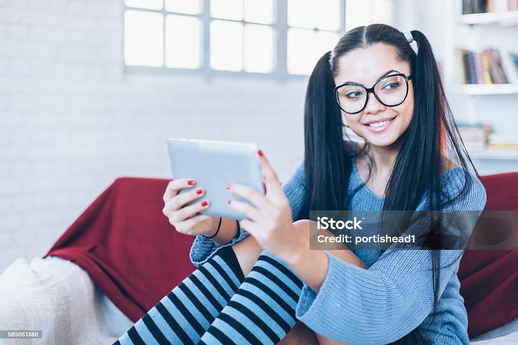 Smiling female student holding digital tablet at home Smiling female student holding digital tablet and sitting on white-burgundy colored sofa. With eyeglasses, wears blue pullover and blue-black knee high long sicks. Looking away. Bookshelfs, windows and brick wall on background. Focus on forerground. Adult Stock Photo
