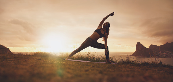 Horizontal shot of healthy female practicing Utthita Parsvakonasana yoga asana. Fitness woman exercising on the cliff during sunset.