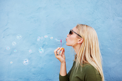 Side view shot of young beautiful female blowing soap bubbles against blue background. Young blond woman having fun.