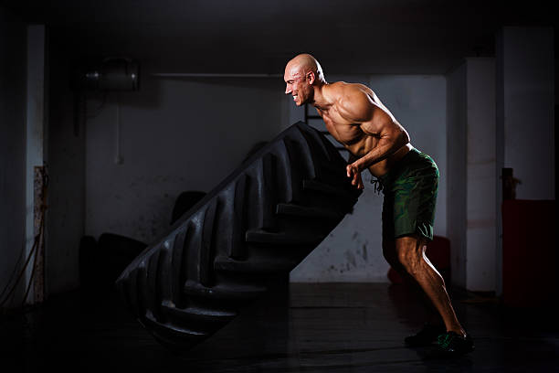 Image of a shirtless muscular bodybuilder doing flipping tire exercise at the gym, Side view.