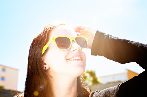 A pretty young brunette in sunglasses stands outdoors looking up, her hand raised to shield her eyes from the dazzling sun.